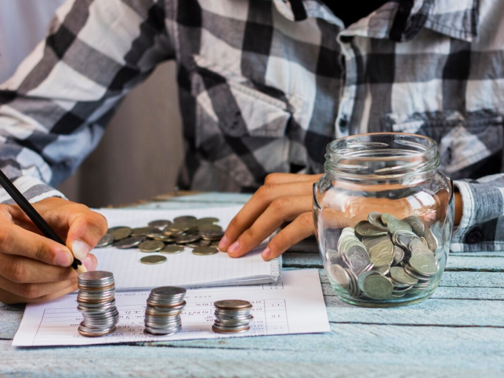 A man sits in front of a pile of change and a jar. He’s organizing and planning his spending strategy using a pen and paper and making individual piles of coins.