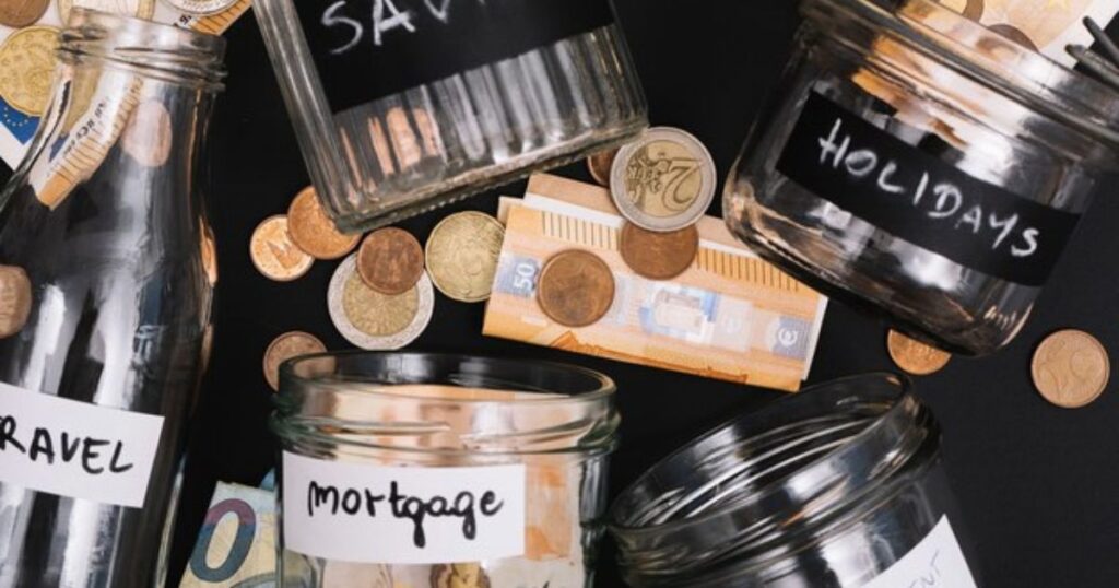  A collection of glass jars with various labels for saving strategies are scattered across a table with a pile of coins and money to be sorted.