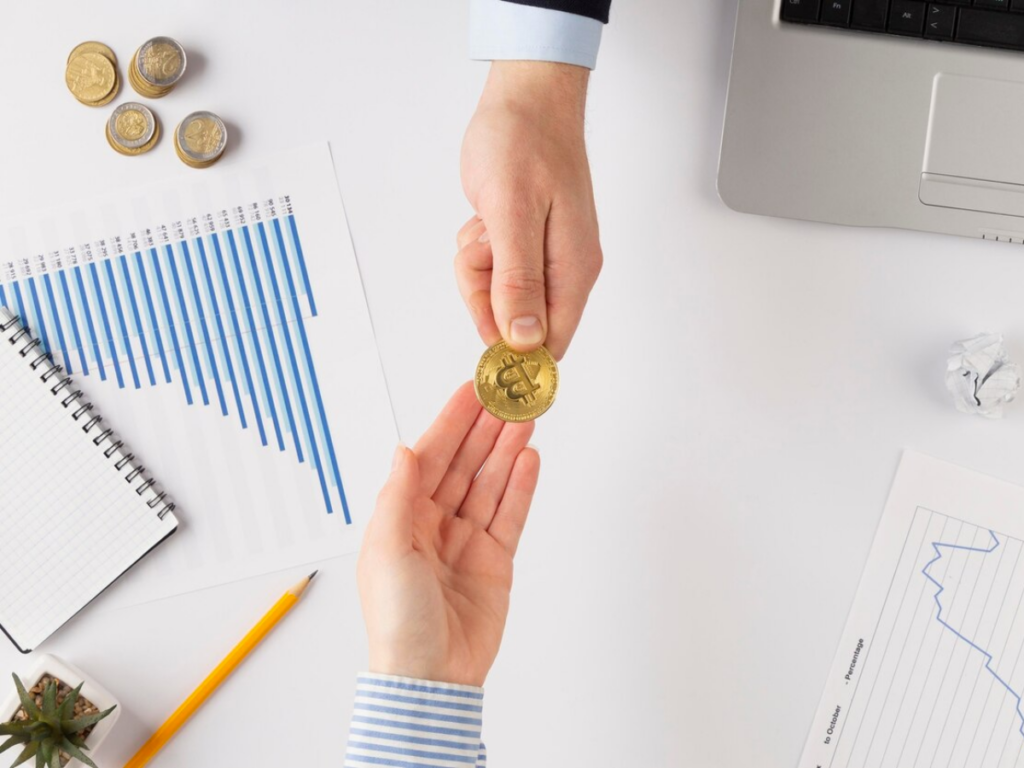 Two hands exchange a Bitcoin over a desk filled with financial charts, a computer, and other coins.