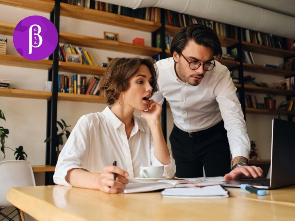 A woman and man work together on taxes with papers spread across a desk and a computer reconciling everything. The woman is happily surprised while the man concentrates. A purple logo for Beta Virtual Assistance.