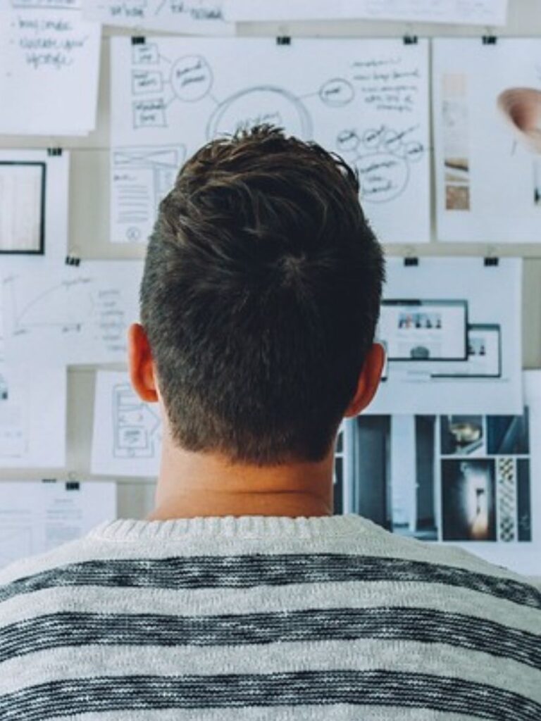 A man stands in front of a wall covered in papers and reports trying to figure out the puzzle