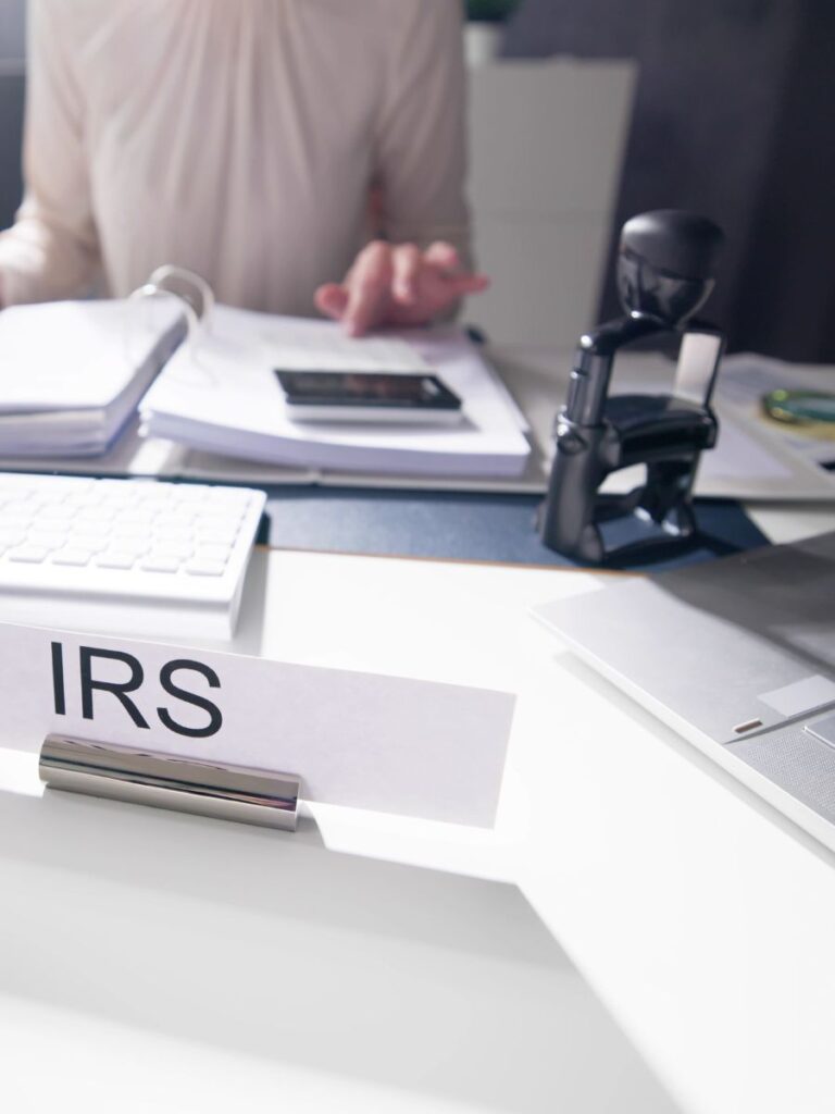 A woman seated at a desk with a binder, calculator, and a sign that says “IRS”.