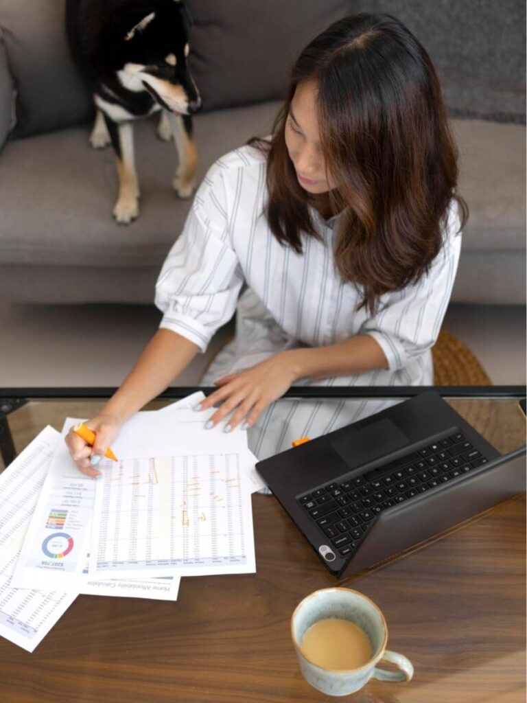 a woman sits on the floor with her computer on a low table and works on her finances by marking up printouts of financial reports. A dog watches from the couch.