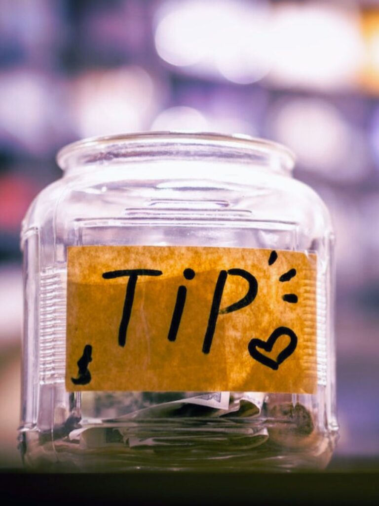 a glass jar with coins inside labeled with a yellow sticky note that says “tip” with a heart drawn next to it