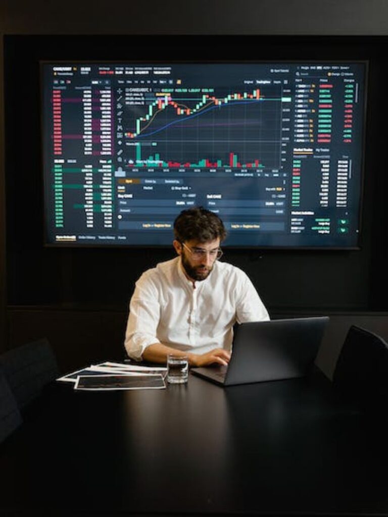 A man works in concentration on his computer; behind him is a large view of a screen open to a crypto exchange and showing trades for a coin