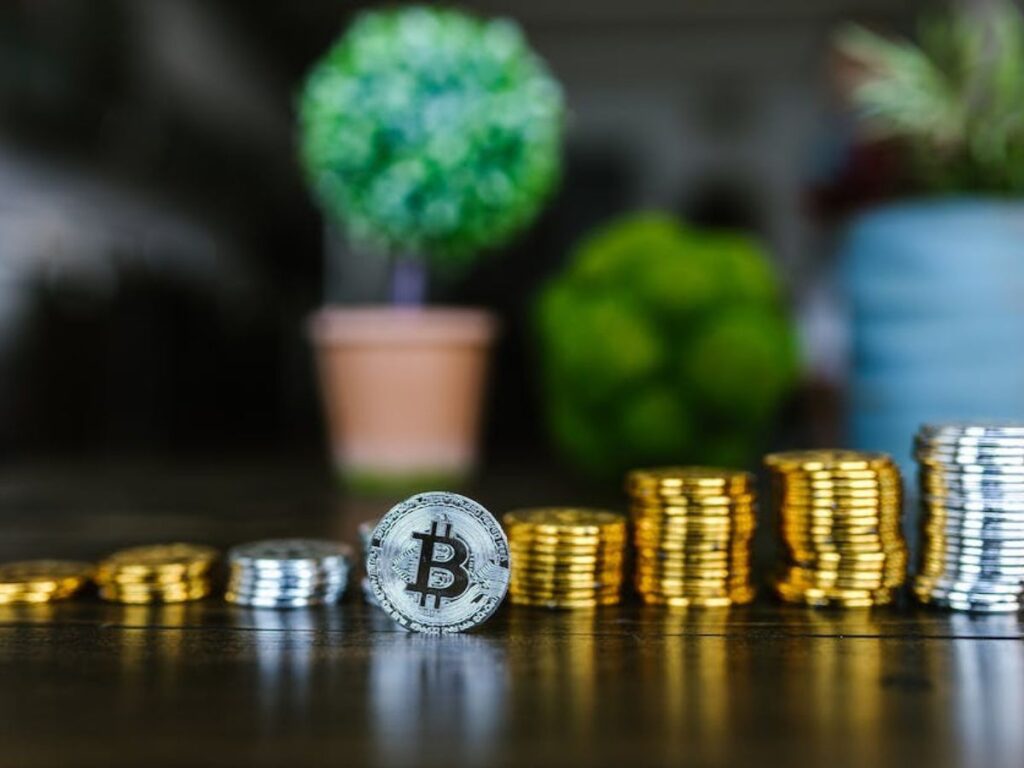 Piles of silver and gold coins growing progressively larger. A bitcoin in front and blurred trees behind.