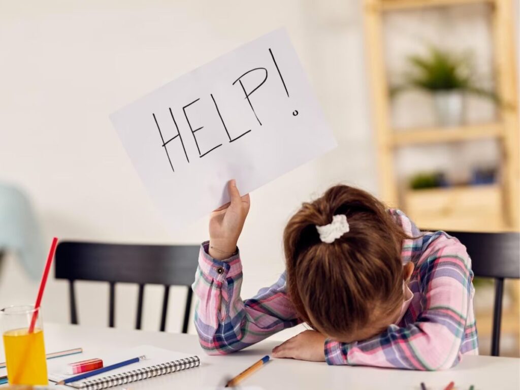 elementary student with her head down on a table surrounded by pencils and school supplies raises a sign over her head that says “Help!”