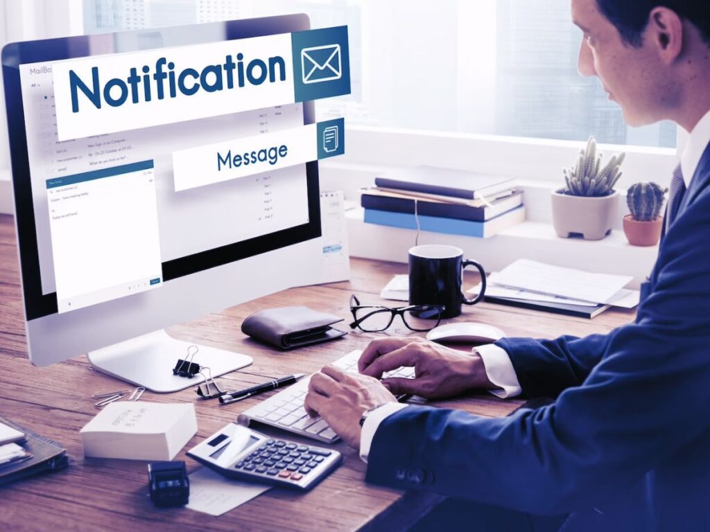 A man working at his computer flashing notifications and alerts for email. Many items clutter his desk relating to work like books, calculator, wallet, glasses.